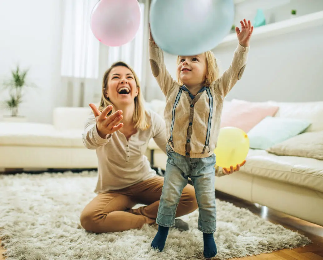 A small boy and his nanny are playing with balloons and smiling. They are on a rug in a stylish living area with a couch behind them