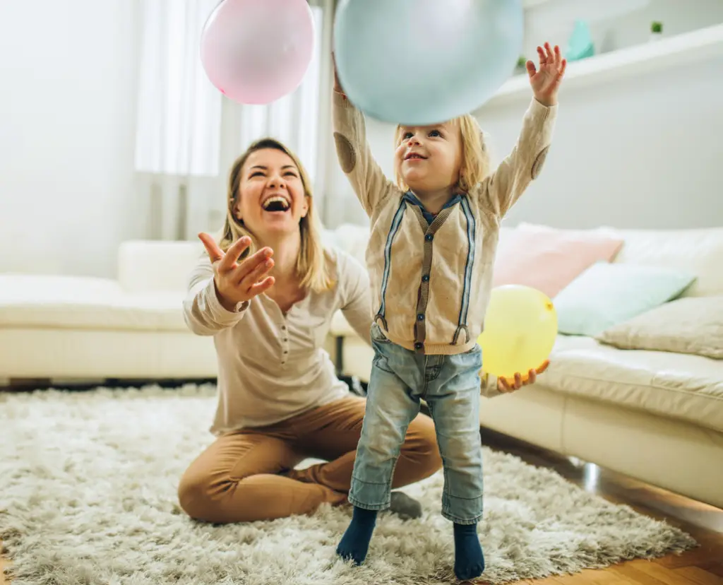 A small boy and his nanny are playing with balloons and smiling. They are on a rug in a stylish living area with a couch behind them